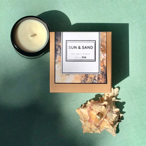 Sun + Sand soy wax candle with its box and a seashell styled on a green textured background.